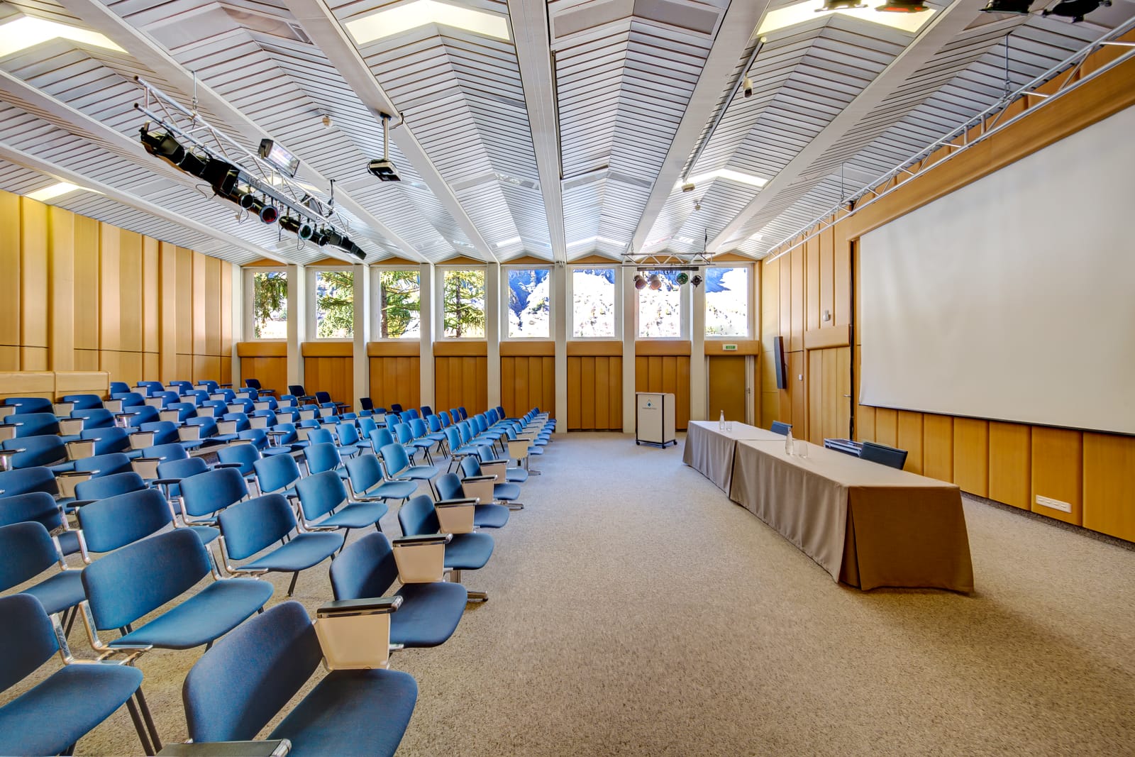 L’aula de la clinique : des rangées de sièges bleus, une table d’orateurs, un écran et un plafond à lames blanches