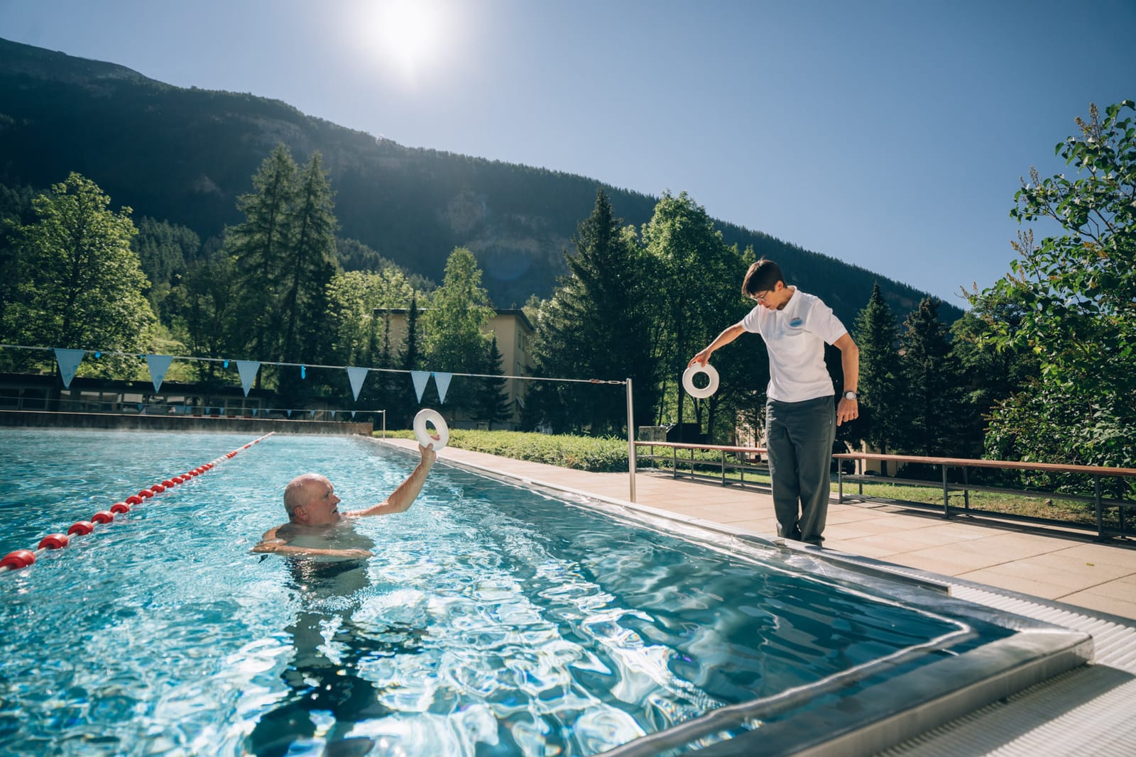 Le bassin extérieur au soleil du matin : une personne nage, un thérapeute longe le bord, sapins et montagne derrière