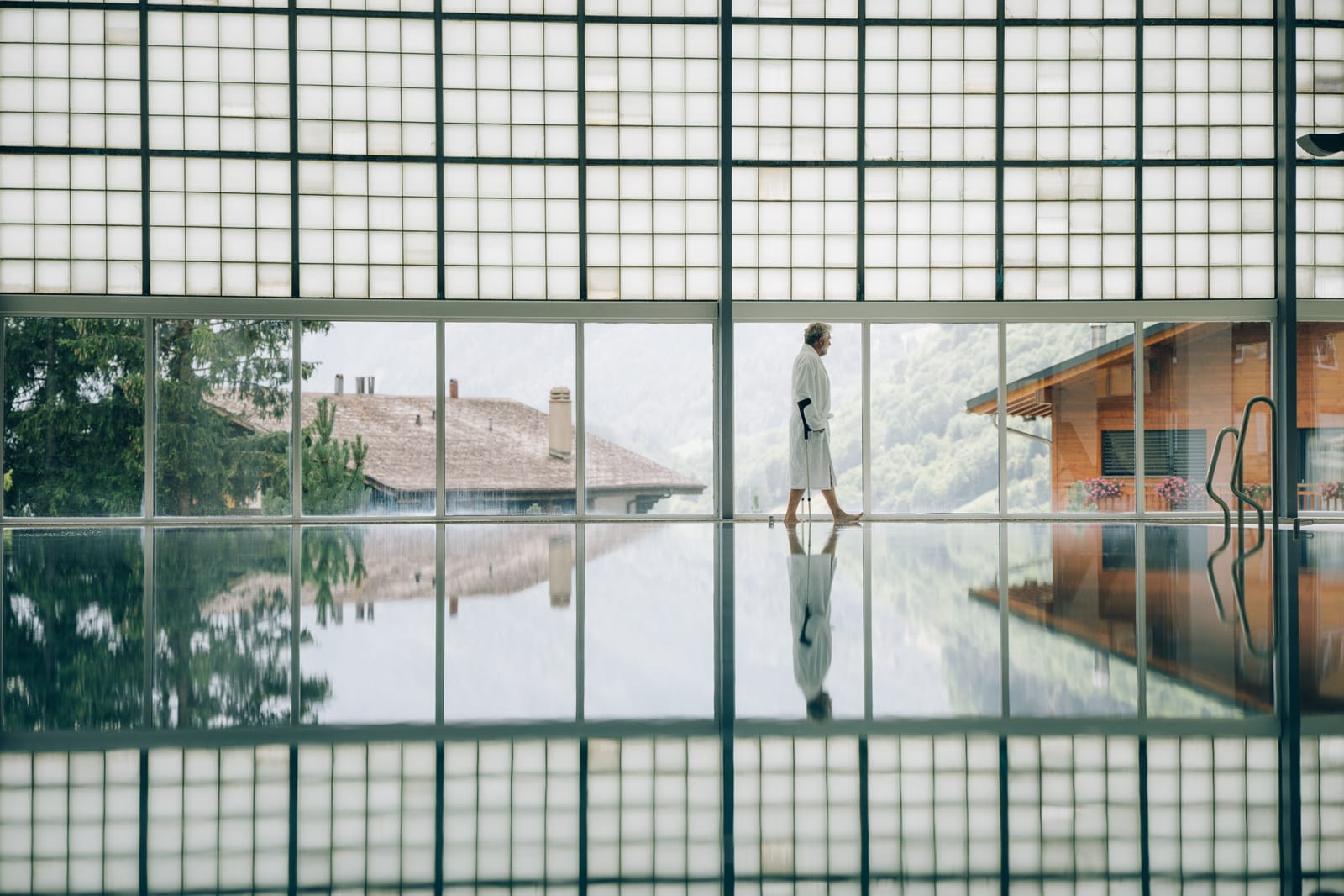 Le bassin thermal intérieur derrière la grande façade vitrée : une personne en peignoir longe le bord, les chalets et la montagne se reflètent dans l’eau