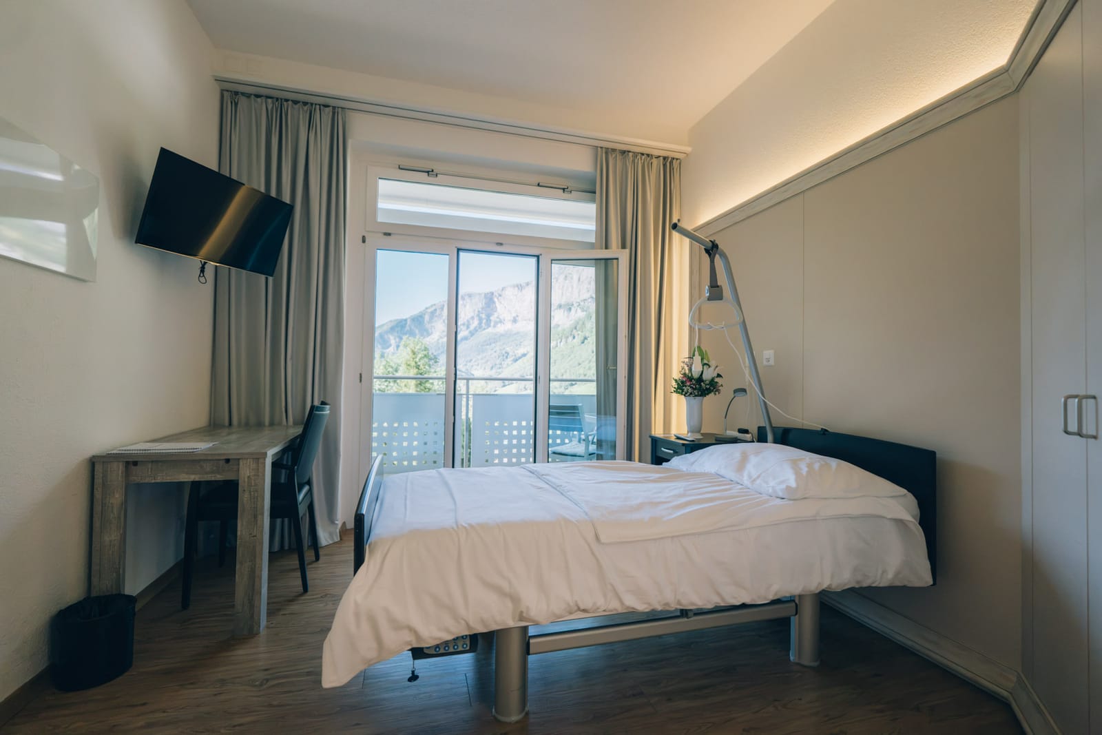 Une chambre de patient : lit, bureau et porte-fenêtre ouverte sur le balcon et la montagne