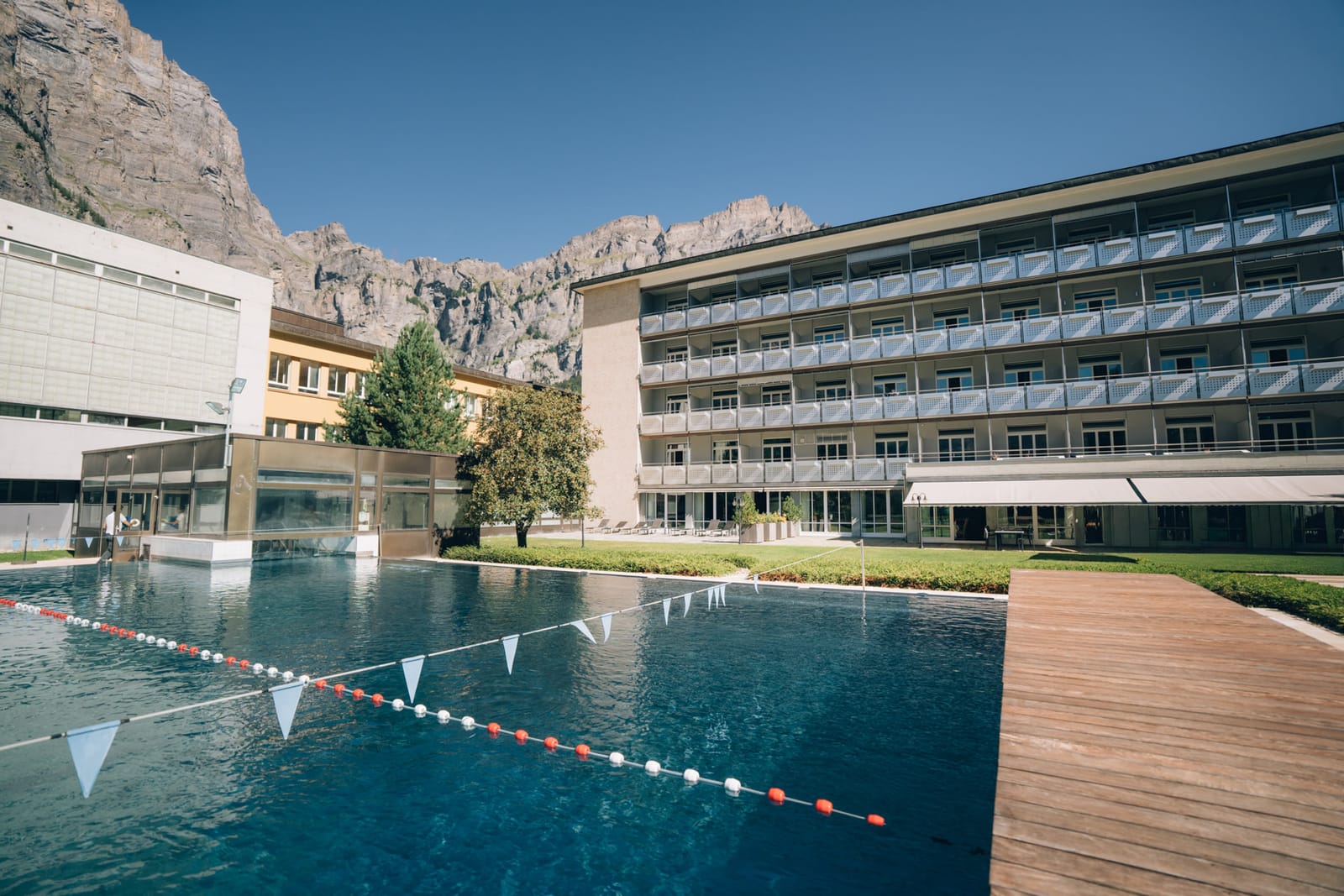 Le bassin extérieur et le bâtiment des chambres de la Leukerbad Clinic un matin d’été, devant les parois rocheuses