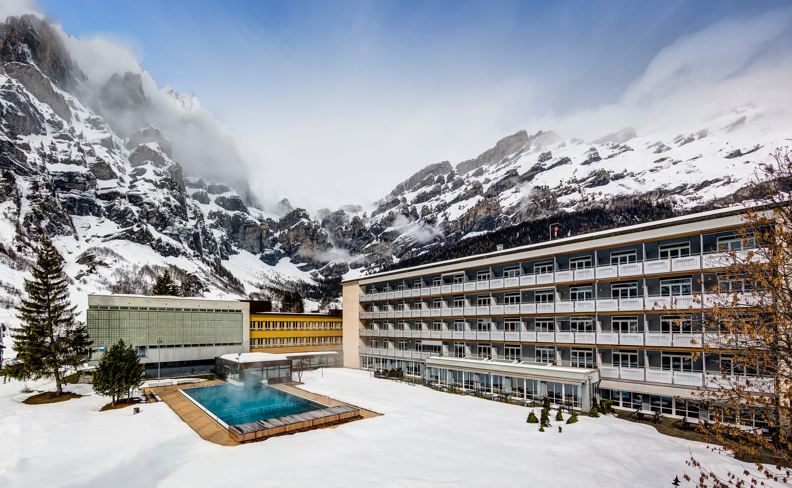 La Leukerbad Clinic en hiver : jardin sous la neige, bassin extérieur fumant, nuages accrochés aux parois