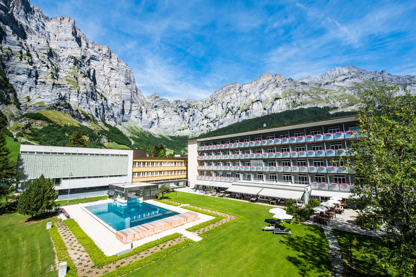 La Leukerbad Clinic en été : le bâtiment des chambres et le bassin extérieur sur la pelouse, et les parois de la Gemmi sous un ciel bleu