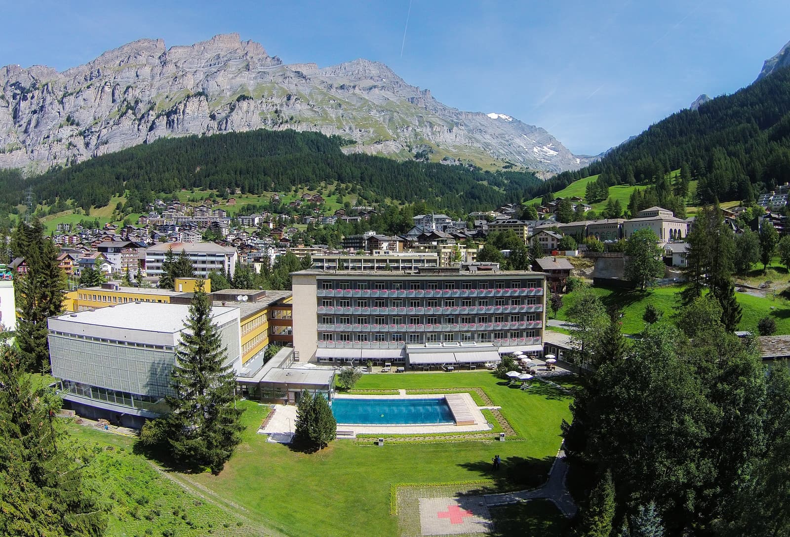 Vue aérienne : la Leukerbad Clinic, son jardin et son bassin extérieur, puis le village de Loèche-les-Bains et la montagne