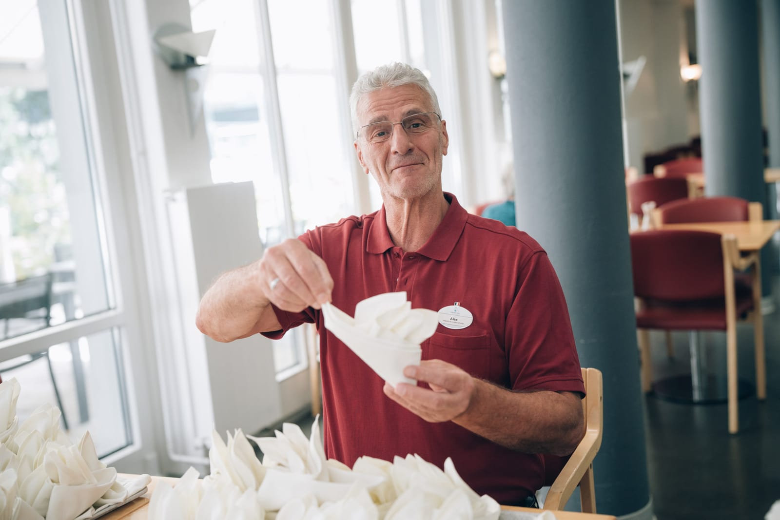 Un collaborateur en polo rouge plie des serviettes blanches dans le restaurant et regarde l’objectif
