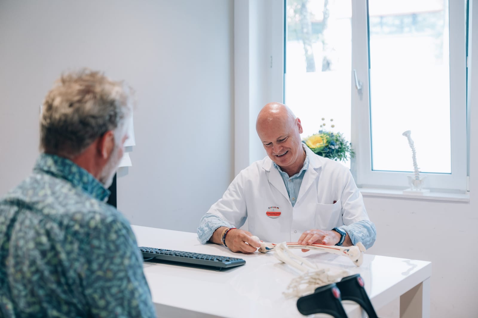 En consultation : un médecin assis à son bureau devant un modèle de squelette, face à un patient vu de dos