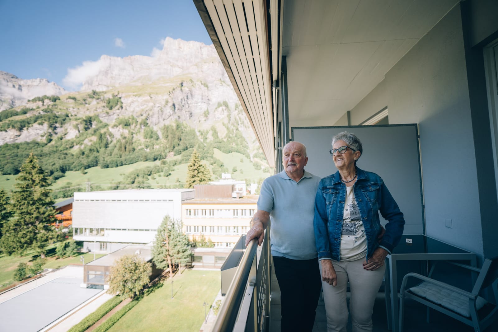 Un couple debout sur le balcon d’une chambre regarde la montagne ; en contrebas, les bâtiments de la clinique