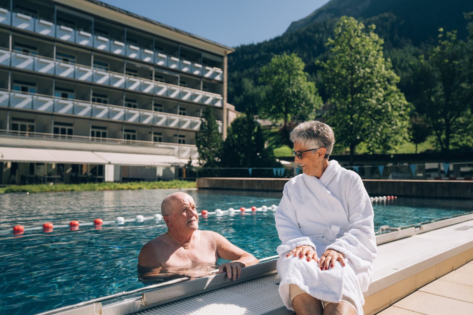 Au bassin extérieur : une femme en peignoir assise au bord parle avec un homme dans l’eau ; derrière, le bâtiment des chambres