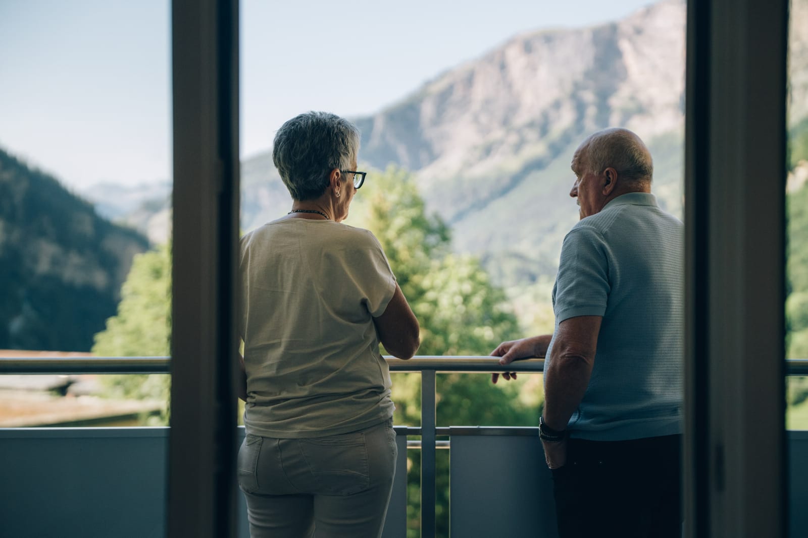 Vu depuis la chambre : deux personnes debout sur le balcon regardent les parois rocheuses et la forêt