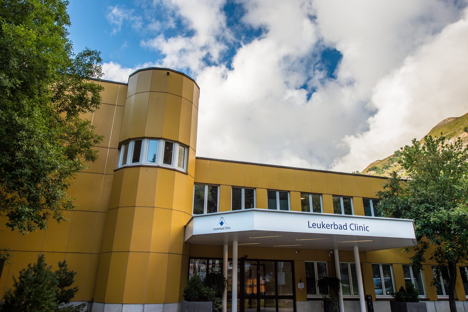 L’entrée principale de la Leukerbad Clinic en été : façade jaune, tour ronde et auvent au nom de la clinique