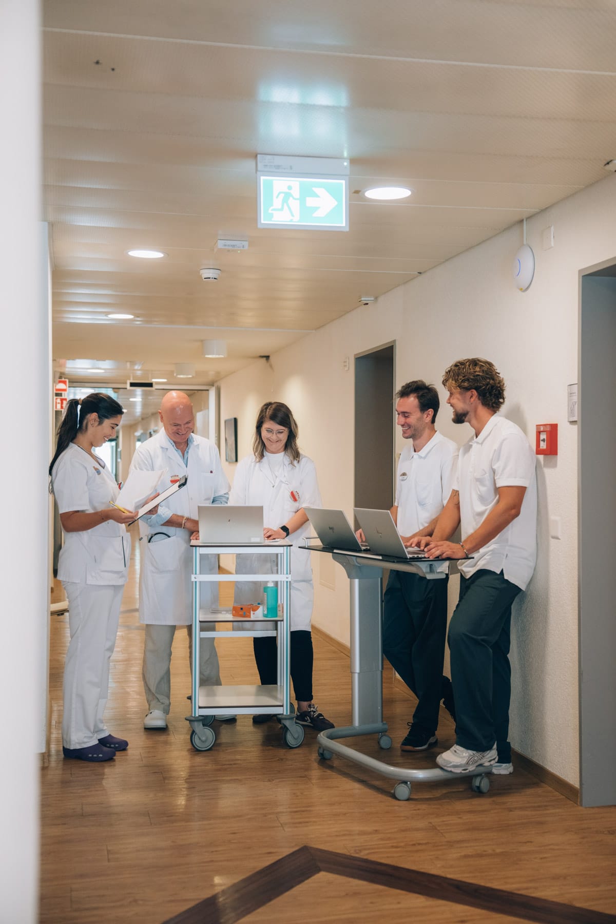 La visite dans le couloir : médecin, soignantes et thérapeutes se concertent autour du chariot de visite et de son ordinateur