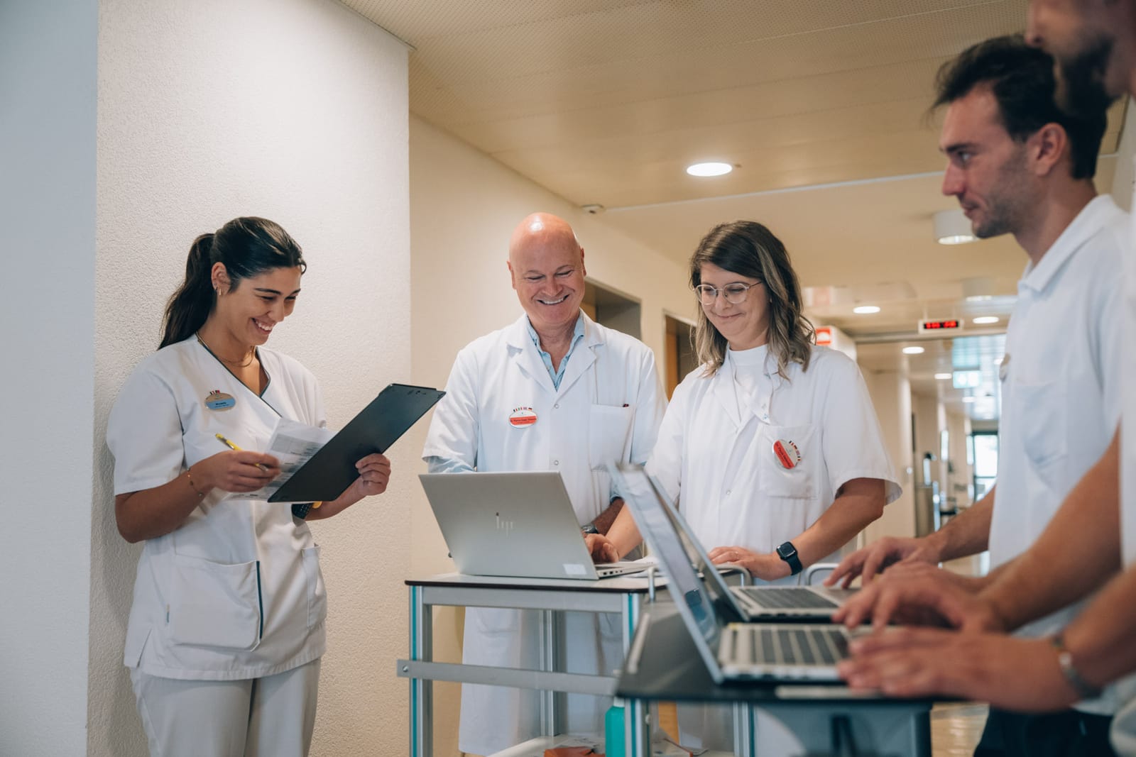 Cinq collègues en tenue blanche se concertent debout autour d’ordinateurs portables et d’une tablette