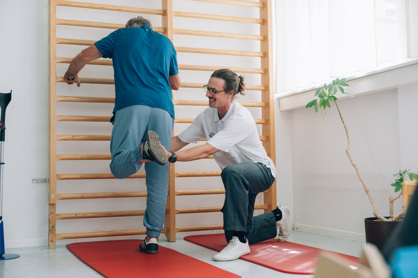 Un patient vu de dos se tient à l’espalier et lève une jambe ; un thérapeute agenouillé guide son pied