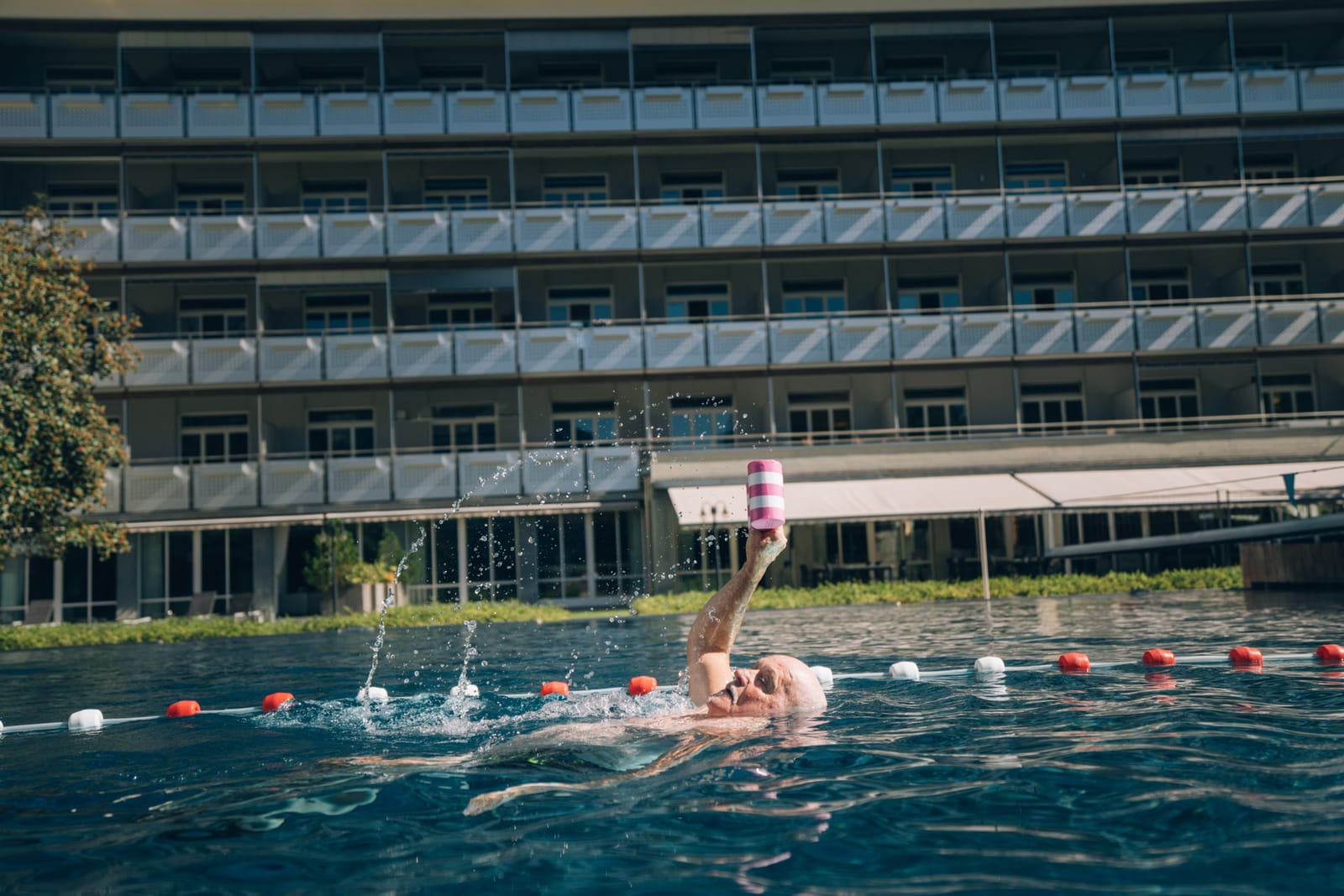 La façade à balcons du bâtiment des chambres au-dessus du bassin extérieur ; dans l’eau, une personne lève un haltère en mousse