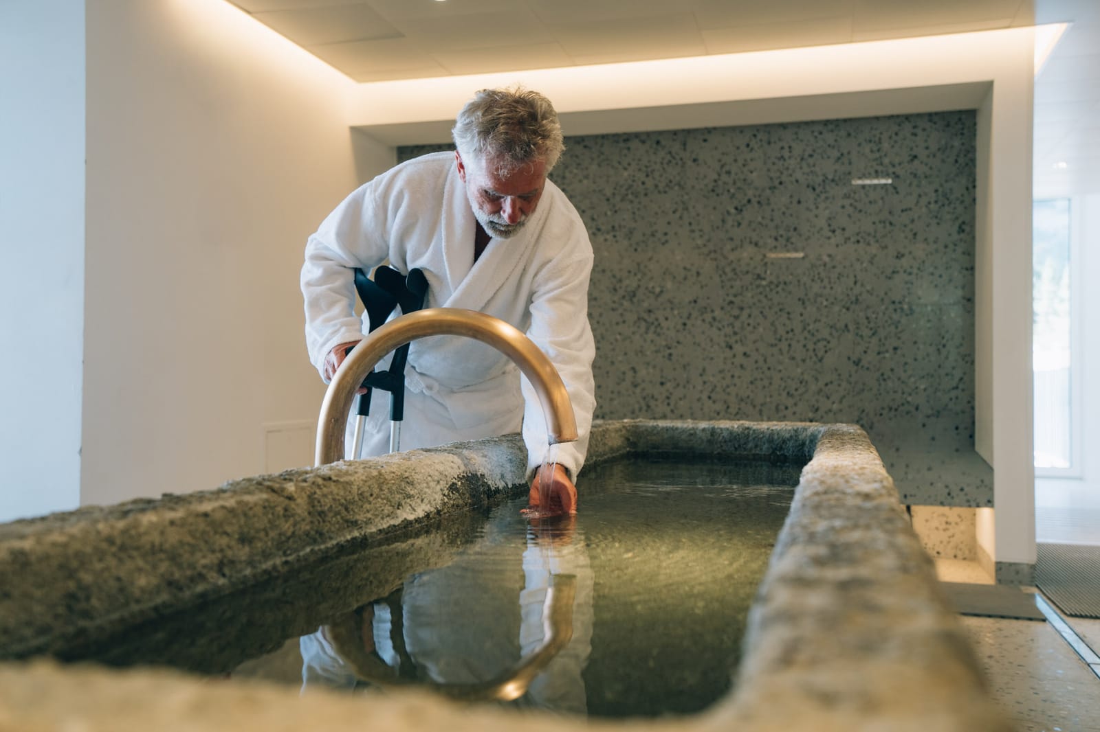 Un homme en peignoir se penche sur le bassin de pierre du spa et plonge la main dans l’eau