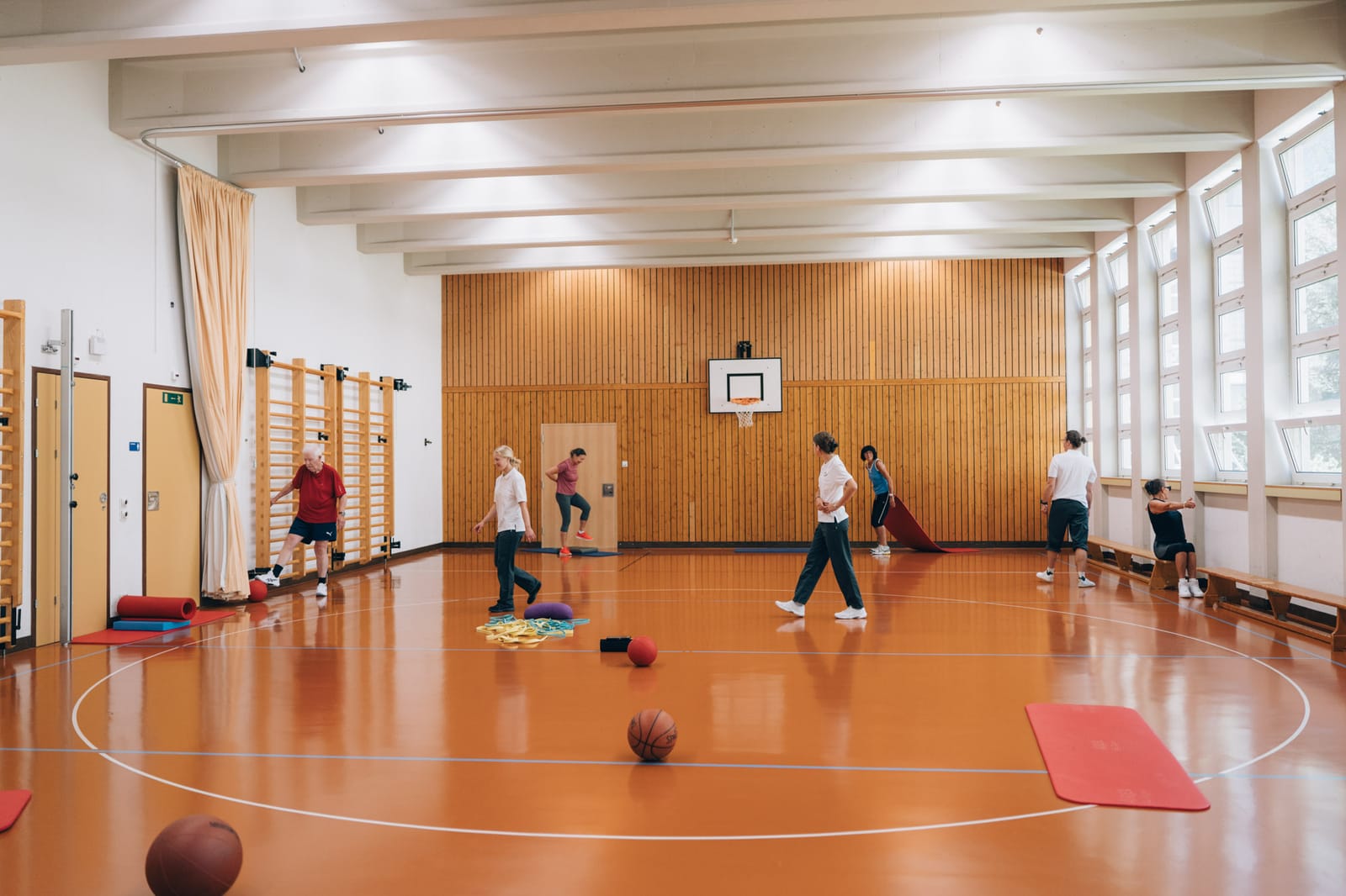 La salle de gymnastique de la clinique vue de loin : un groupe s’exerce avec ballons et tapis, des espaliers au mur