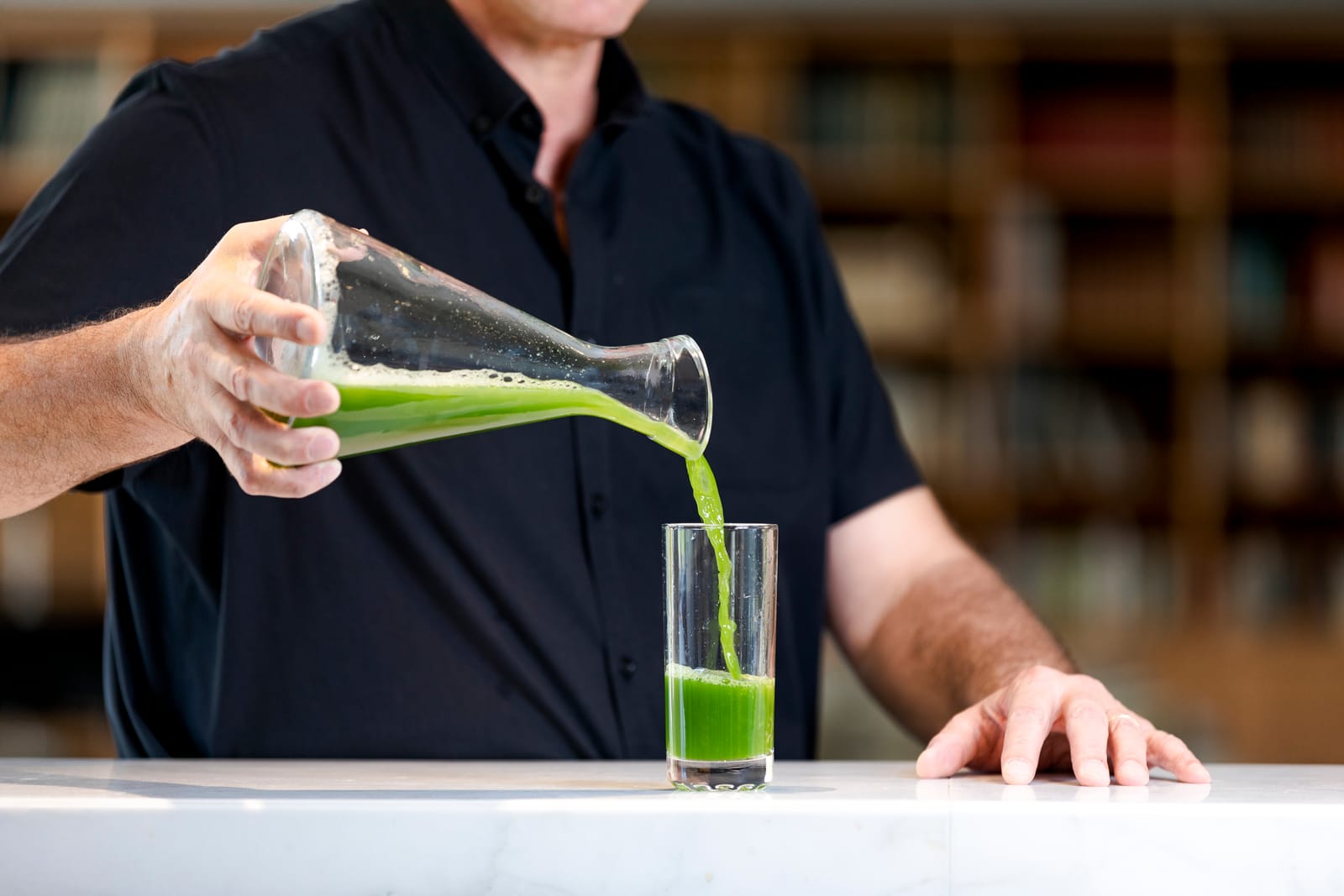 Une main verse un jus de légumes vert d’une carafe dans un verre