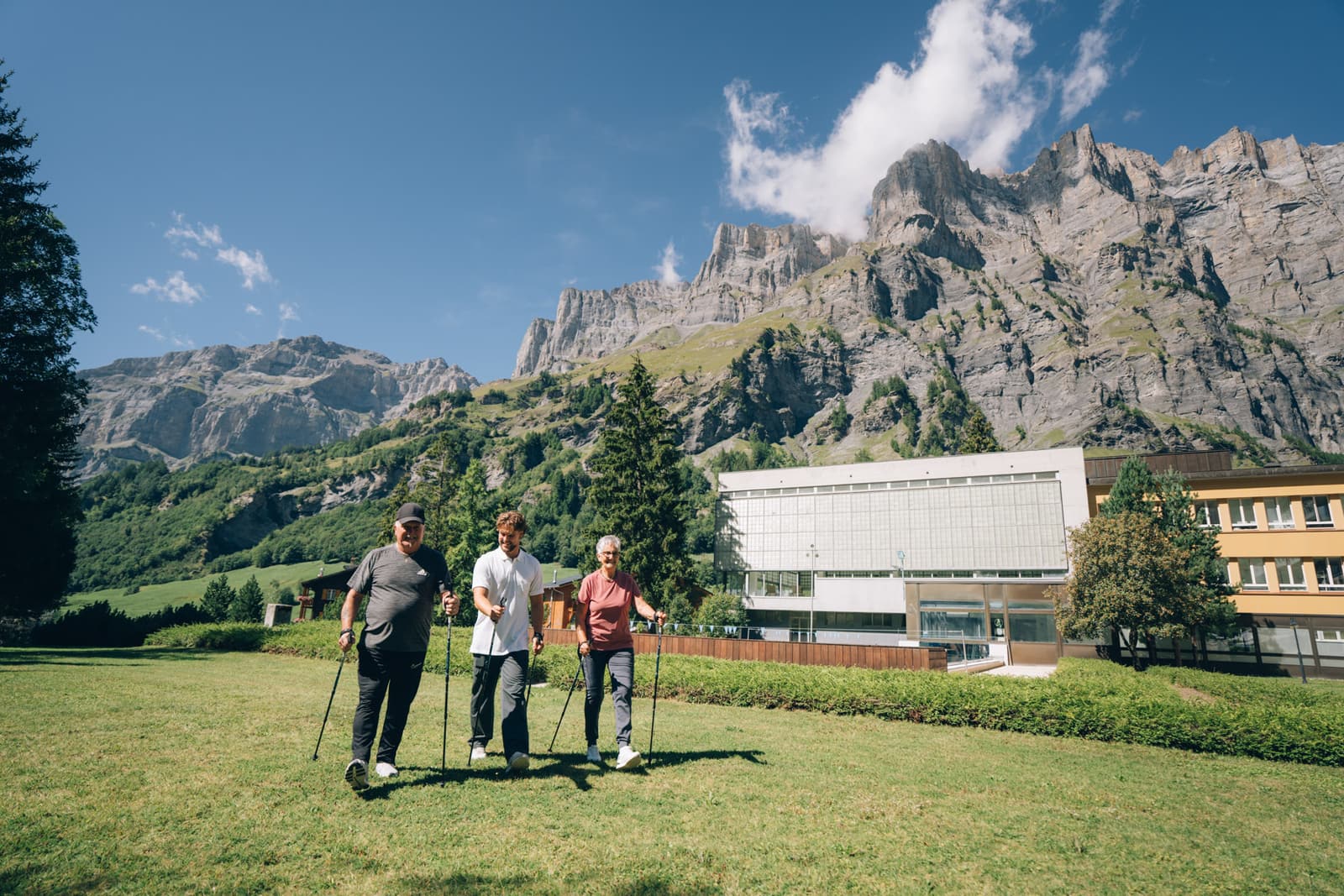 La pelouse devant la Leukerbad Clinic en été, trois marcheurs au loin avec leurs bâtons, et les parois de la Gemmi au-dessus