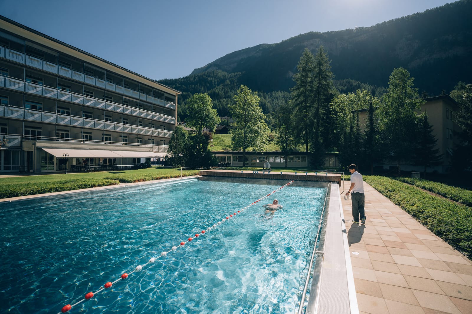 Le bassin extérieur et ses lignes d’eau mènent au bâtiment des chambres ; un thérapeute au bord, une personne dans l’eau