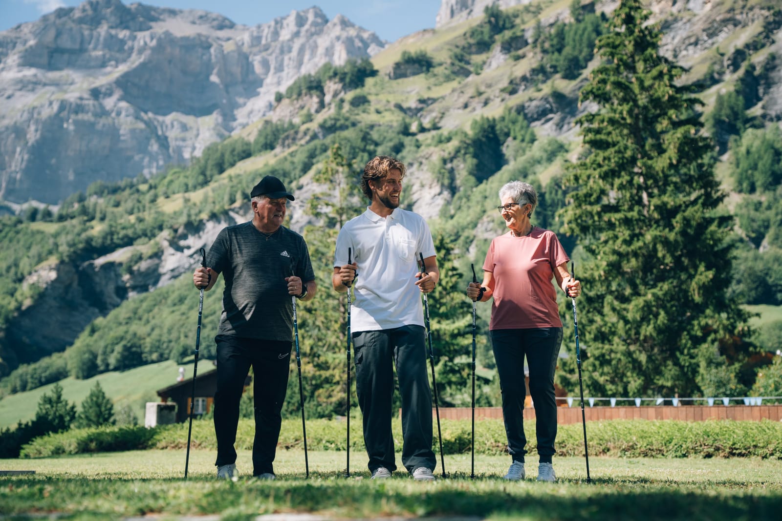 Un collaborateur de la clinique marche avec deux personnes, bâtons en main, sur la pelouse ; derrière, la forêt et les parois