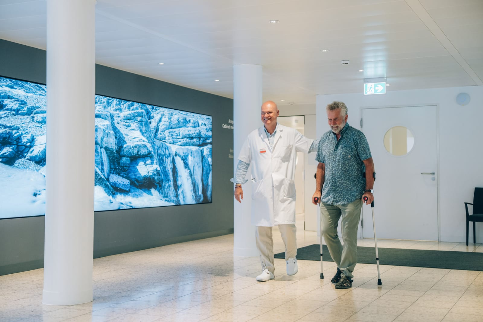 Un médecin accompagne un patient qui marche avec des cannes dans le hall d’entrée ; au mur, une grande image de glacier