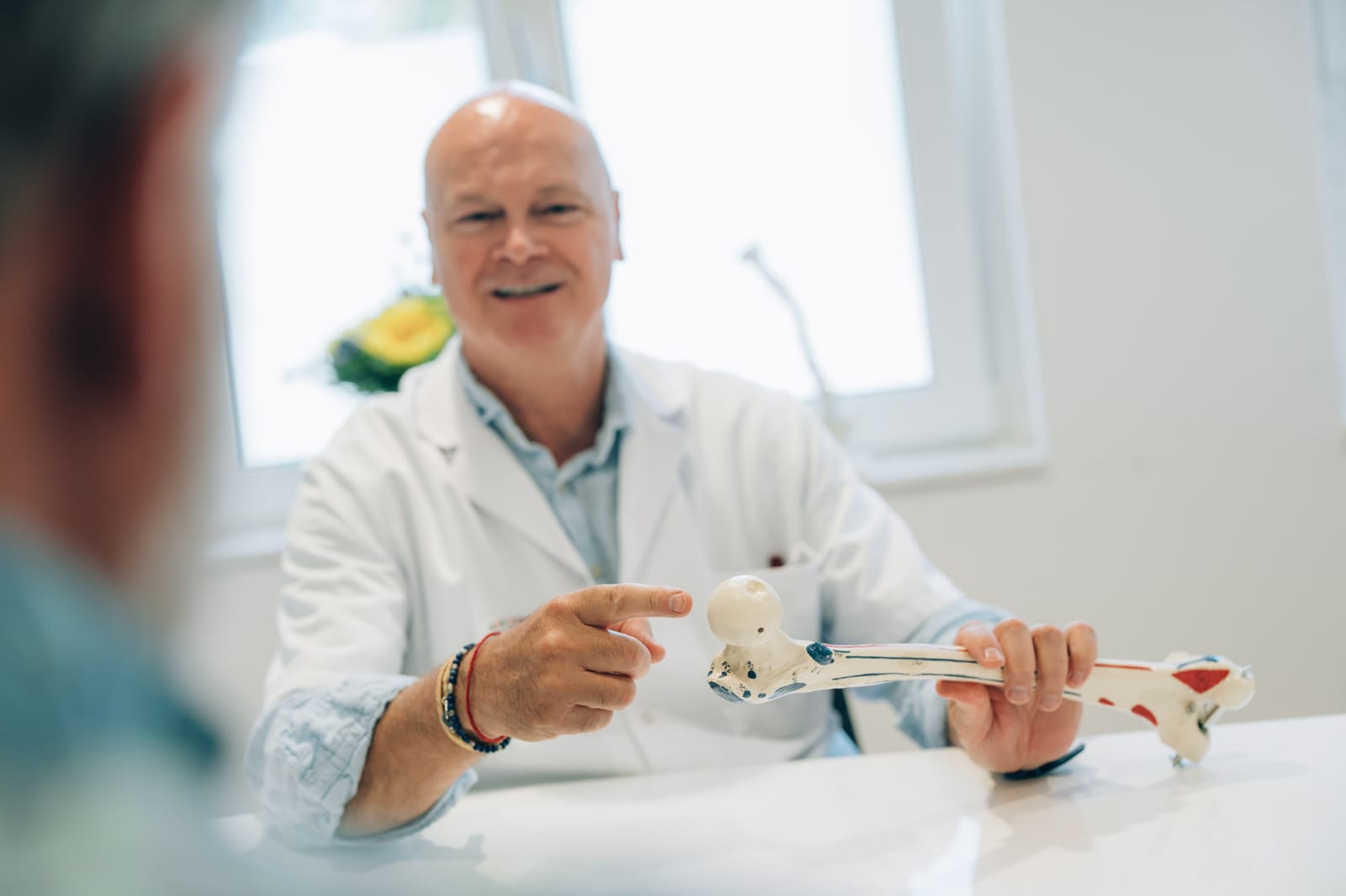 Un médecin en blouse blanche, souriant, explique quelque chose sur un modèle d’os qu’il tient en main