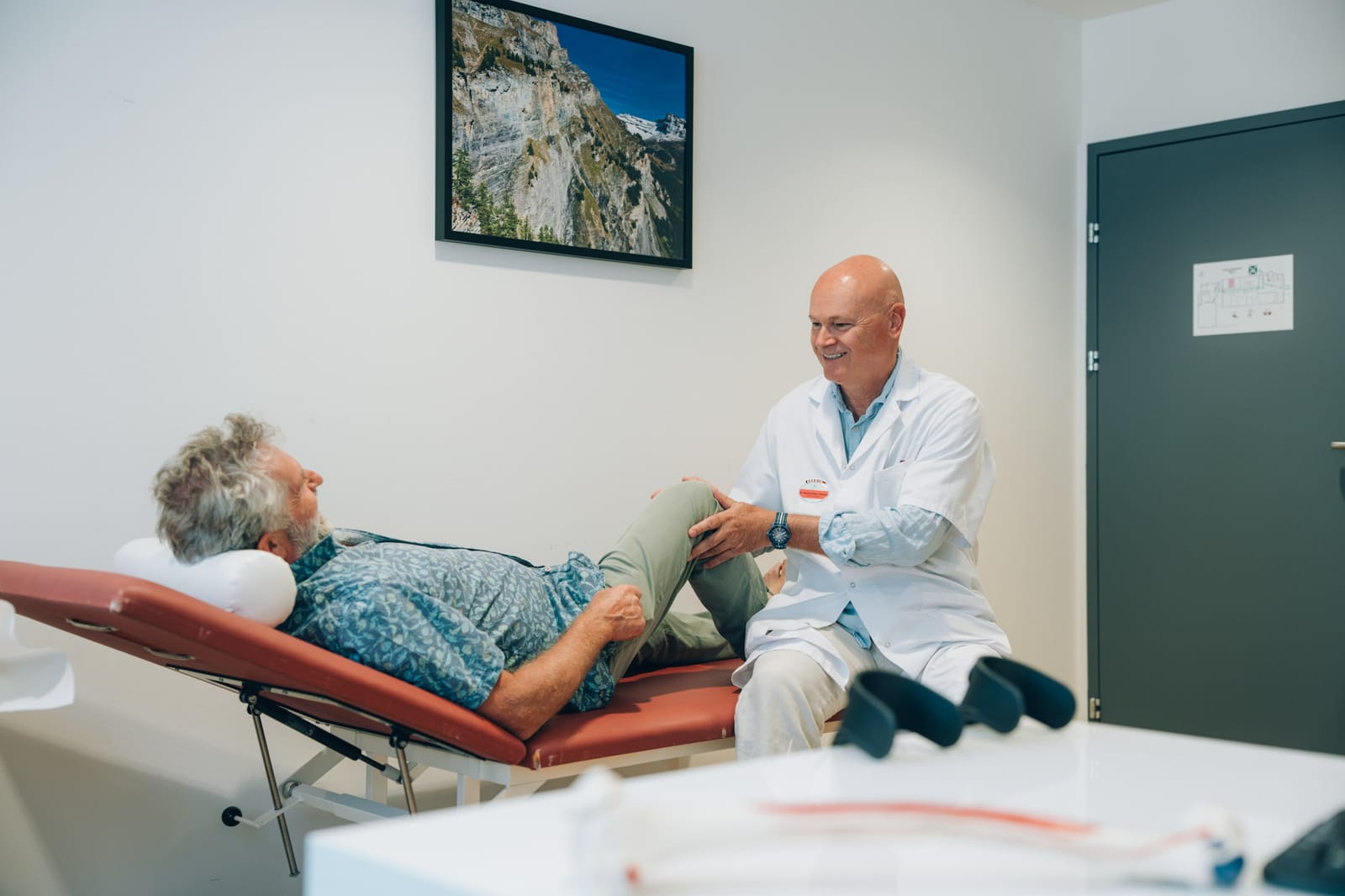 En salle d’examen : un médecin en blouse blanche, souriant, assis près d’un patient allongé sur la table