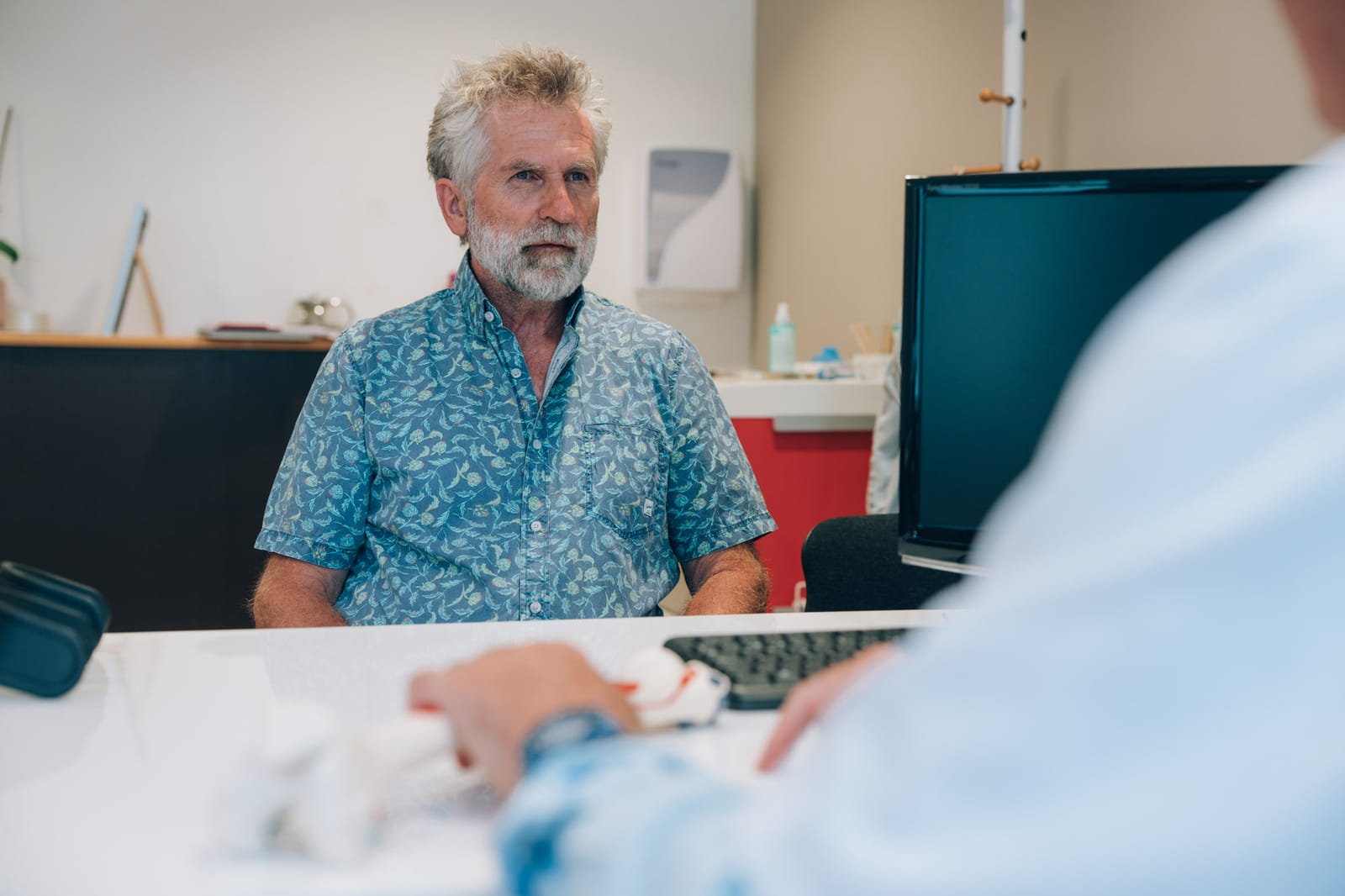 Vu par-dessus l’épaule du médecin : un patient en chemise bleue, assis au bureau du cabinet, écoute