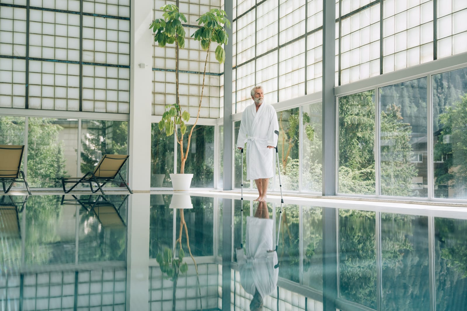 Le bassin thermal intérieur sous la paroi de pavés de verre ; à l’autre bout, une personne en peignoir blanc, chaises longues et plantes