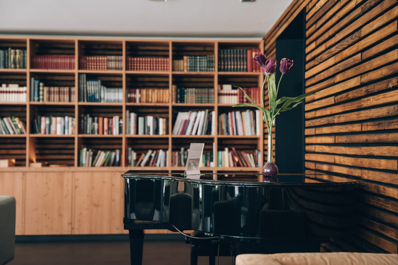 Un piano à queue noir devant le mur de livres ; à gauche, une paroi de lattes de bois et un vase de tulipes