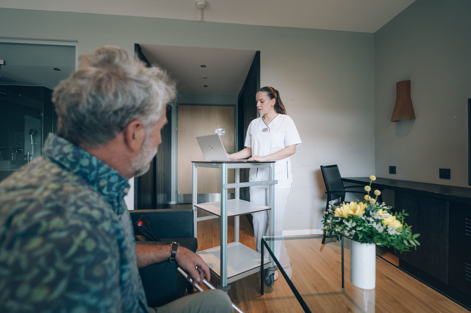 Un patient vu de dos s’adresse, dans sa chambre, à une soignante debout au chariot de visite ; des fleurs jaunes sur la table