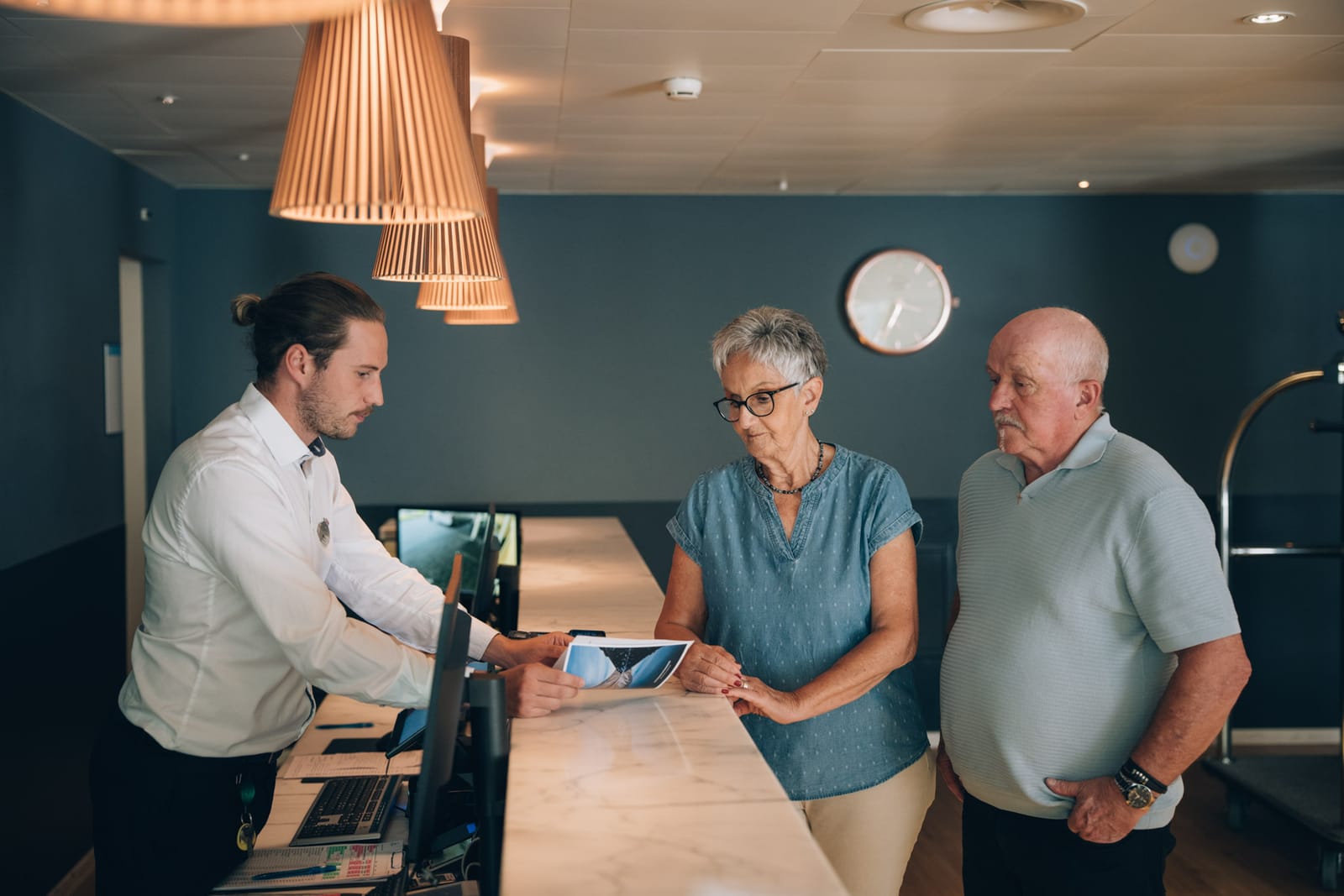 À la réception, un collaborateur montre quelque chose sur une tablette à un couple ; suspensions en bois et horloge au mur