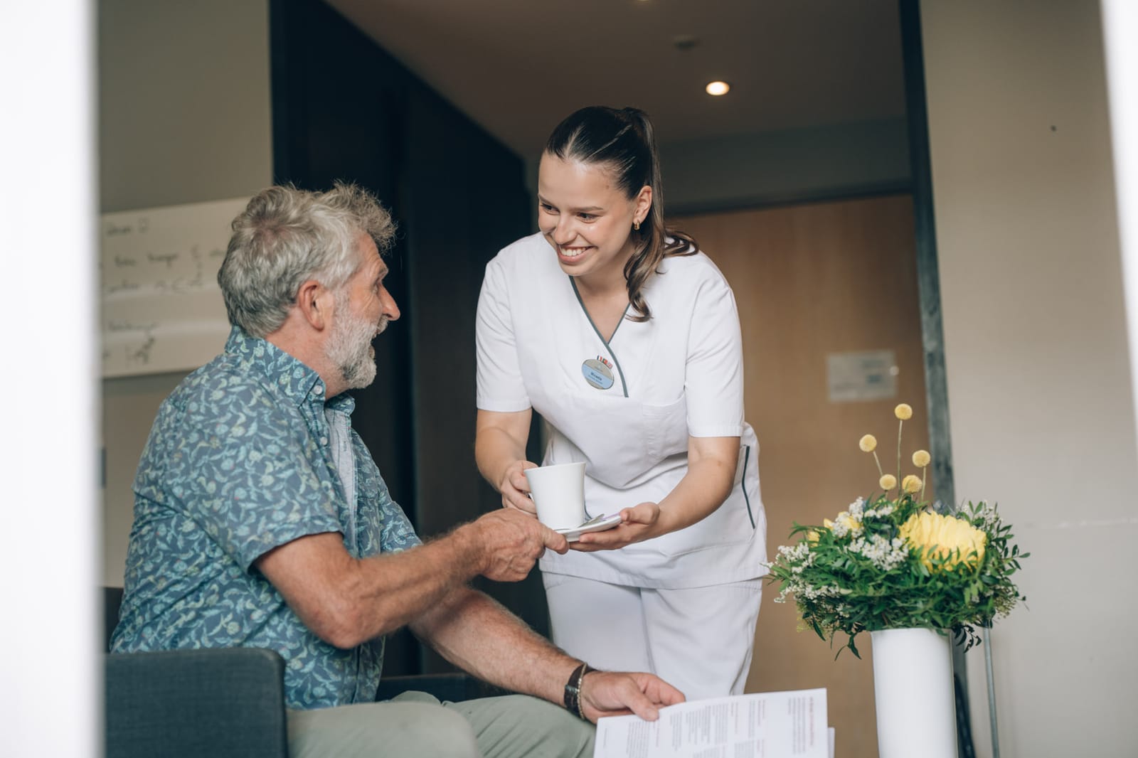 Une soignante souriante tend une tasse à un patient assis ; sur la table, un bouquet de fleurs