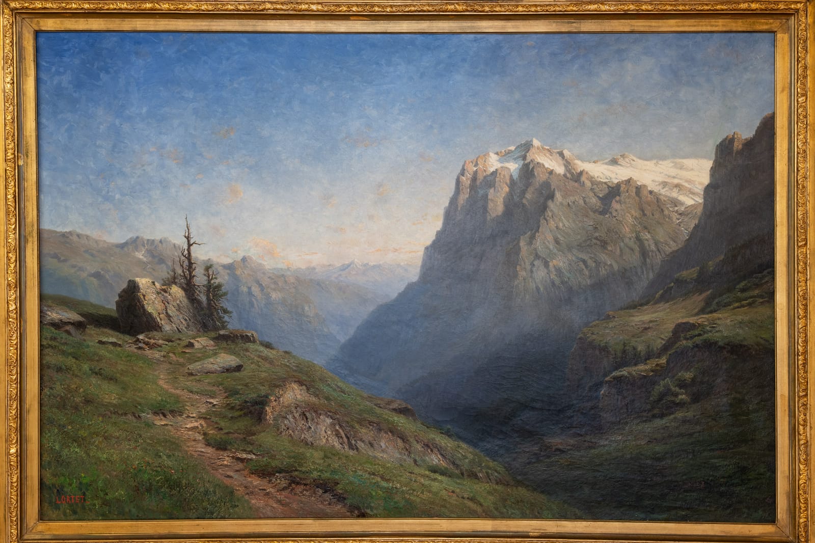 Un tableau de la collection de la clinique : un sentier de montagne au-dessus d’une vallée, face à un sommet rocheux enneigé dans la lumière