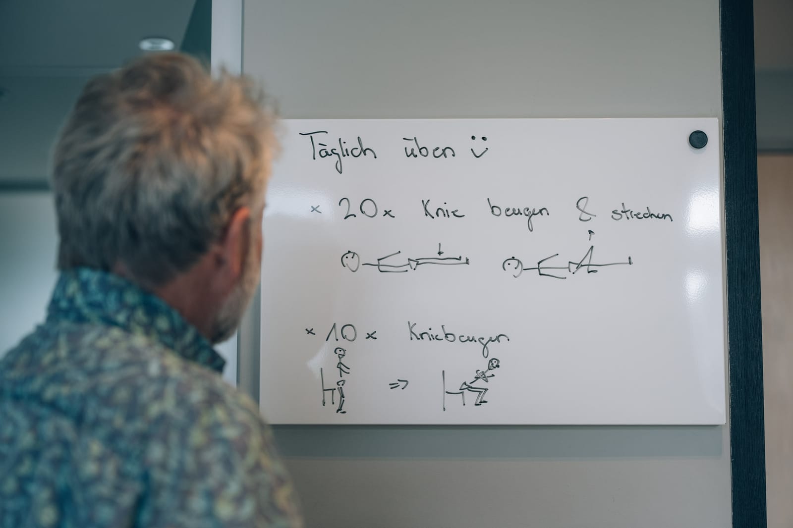 Un patient vu de dos lit, dans sa chambre, le tableau de ses exercices quotidiens : vingt flexions du genou, dix flexions sur jambes