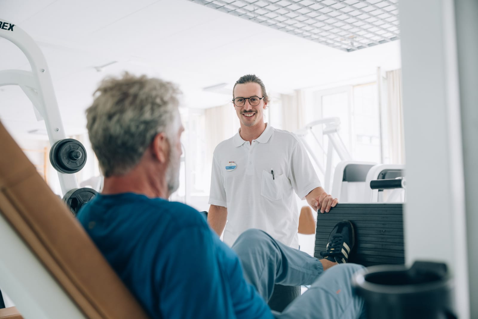 En salle d’entraînement : un thérapeute souriant se tient près d’un patient assis à un appareil