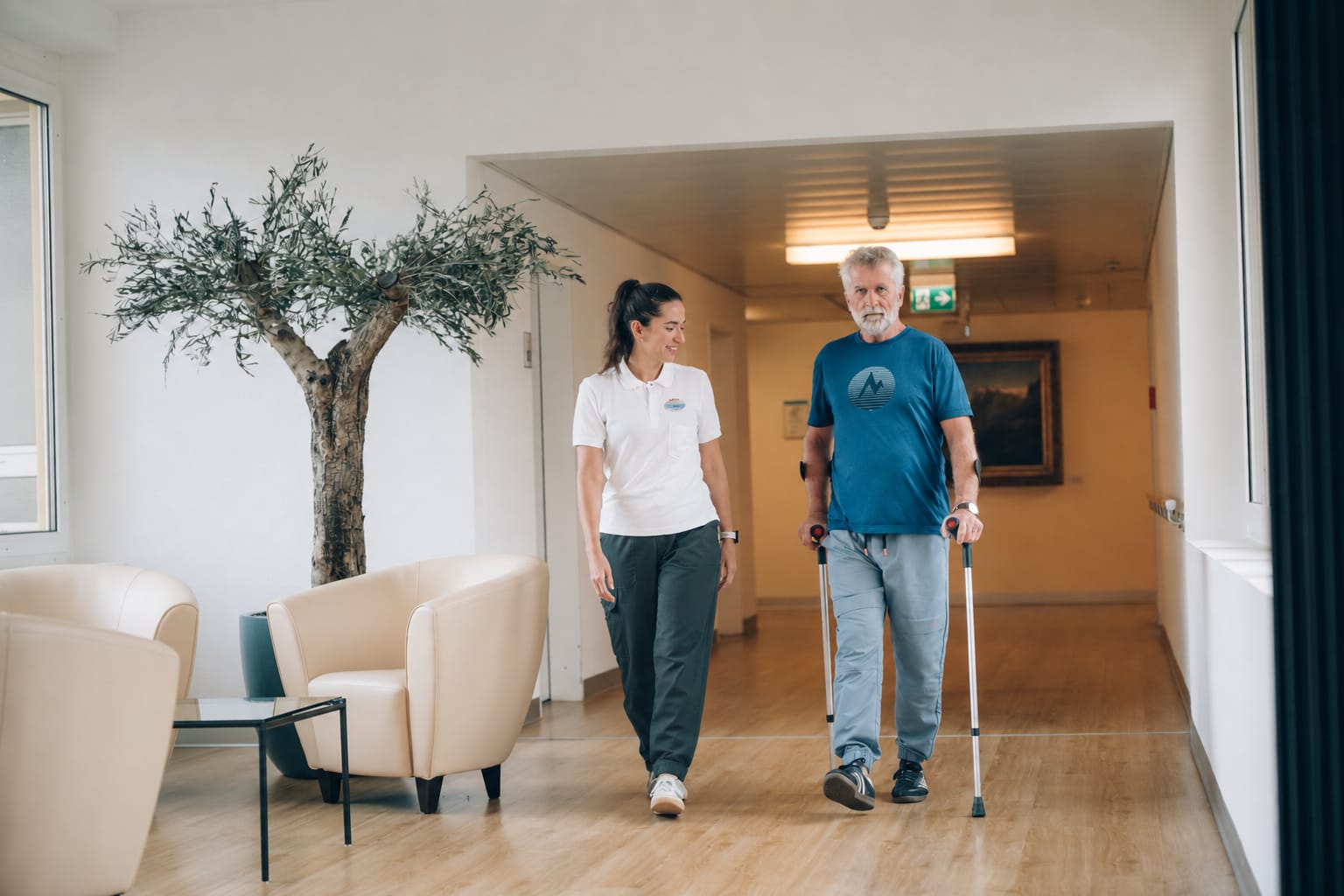 Dans le couloir clair, un patient avance avec deux cannes à côté d’une thérapeute souriante, devant un olivier et des fauteuils