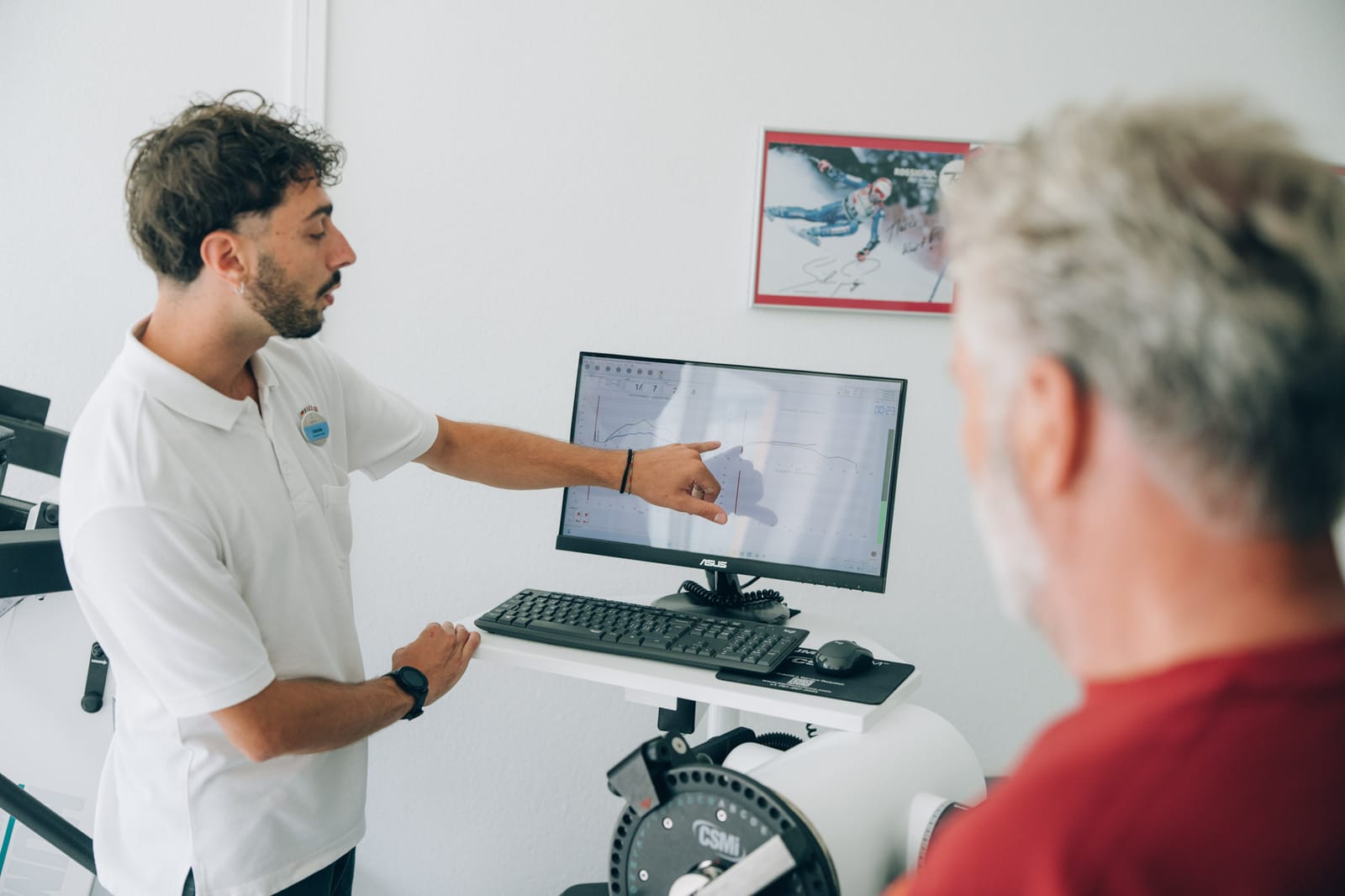 Un thérapeute montre du doigt une courbe de mesure sur l’écran d’un appareil de test de force ; au premier plan, flou, un patient